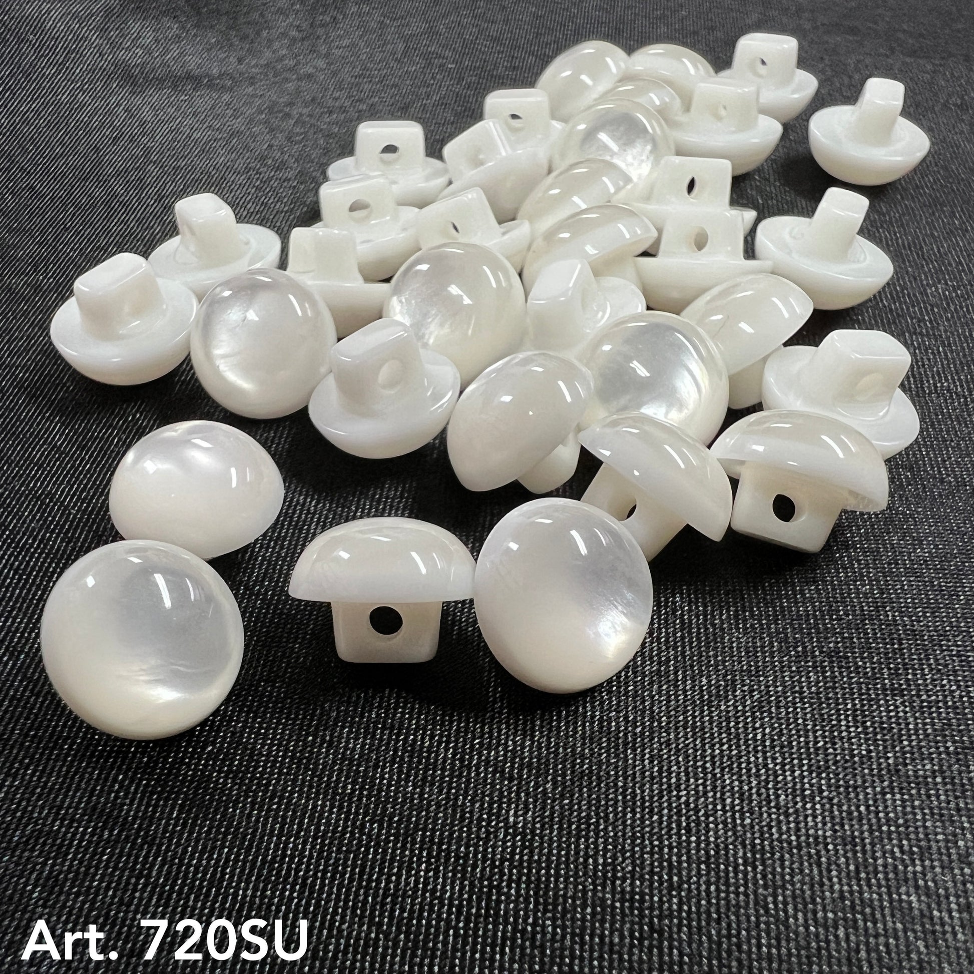 Buttonology Inc white pearl round shank buttons for garments scattered on dark denim fabric