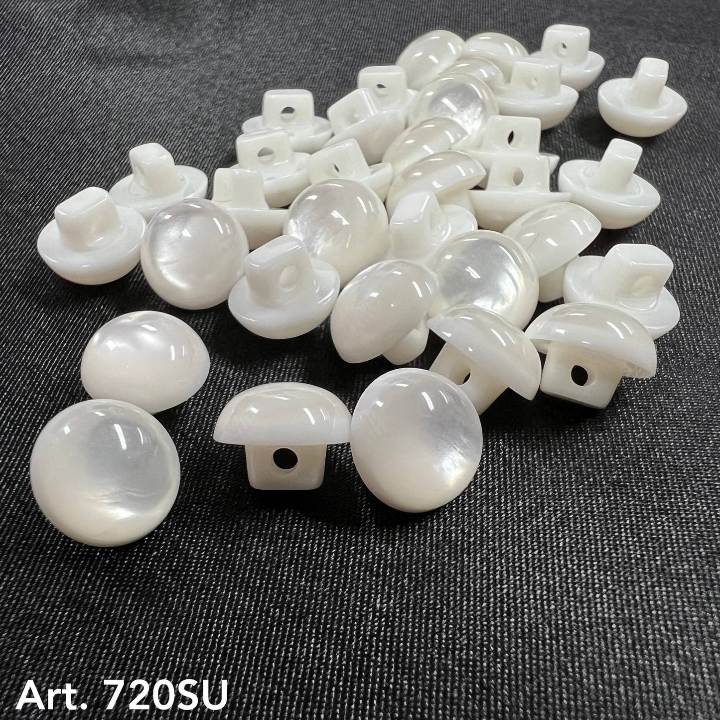 Buttonology Inc white pearl round shank buttons for garments scattered on dark denim fabric