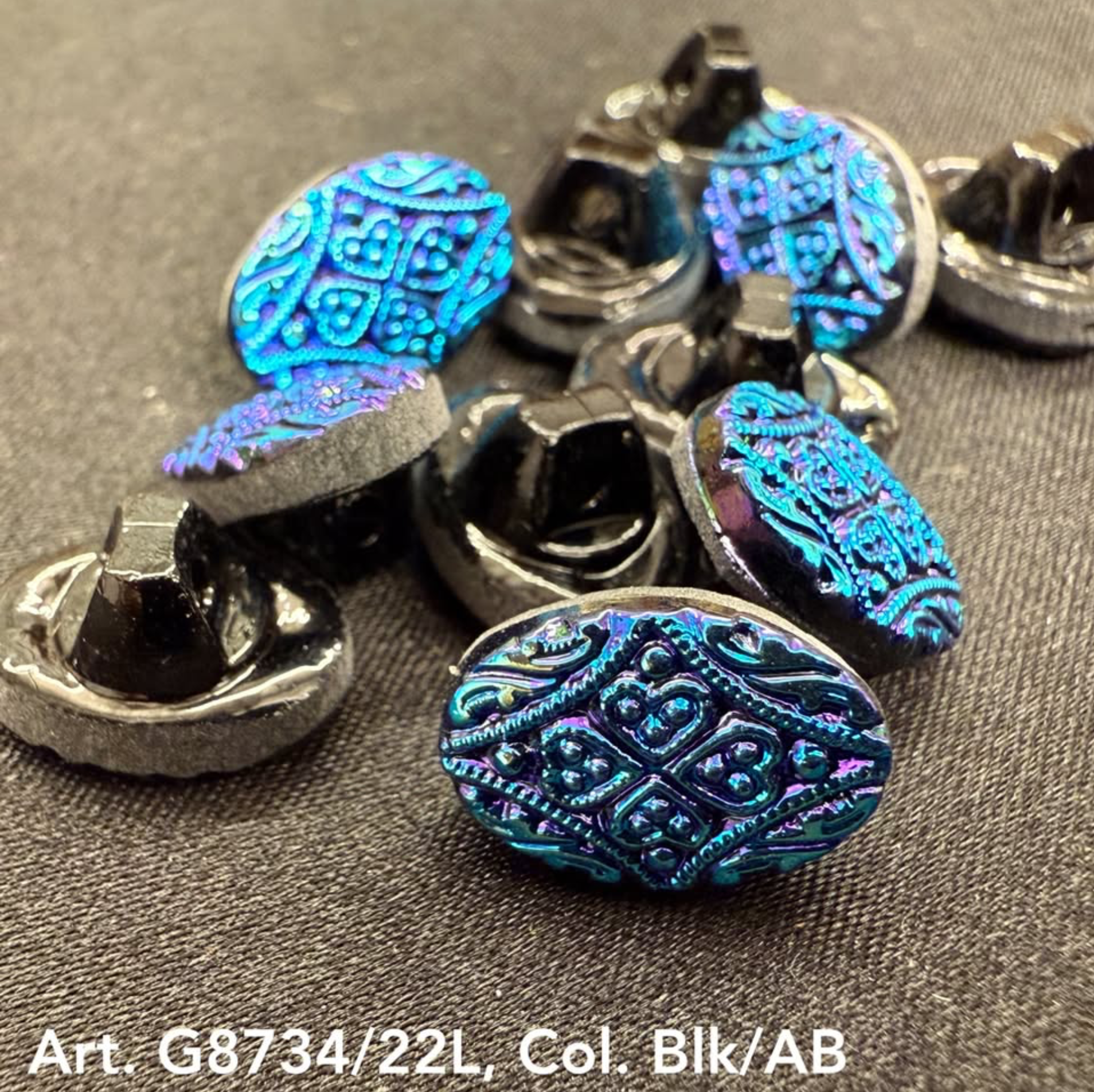 Blue decorative garment button with intricate pattern from Buttonology Inc on a dark fabric background