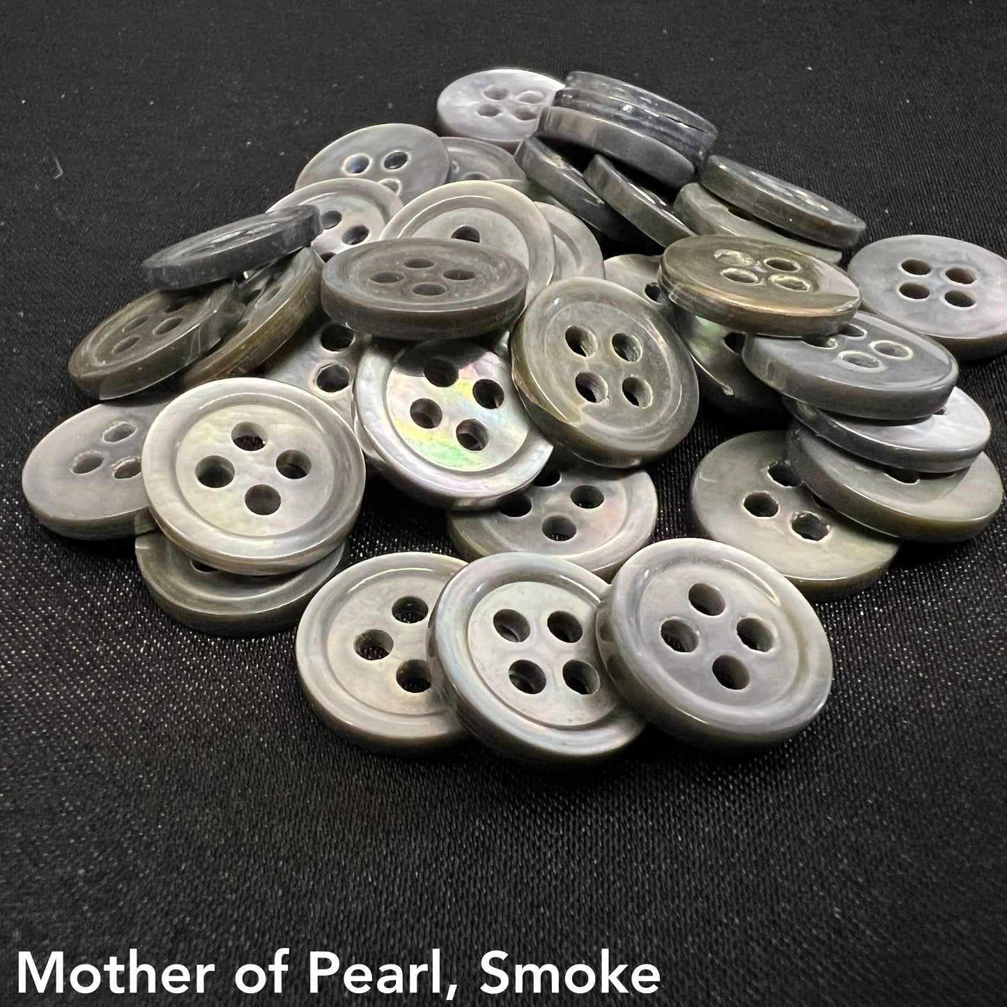 Pile of gray and black mother of pearl buttons with four holes by Buttonology Inc on black fabric