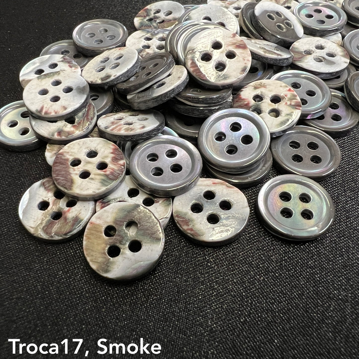 Troca shell buttons for garments by Buttonology Inc in smoke color with four holes on black fabric background