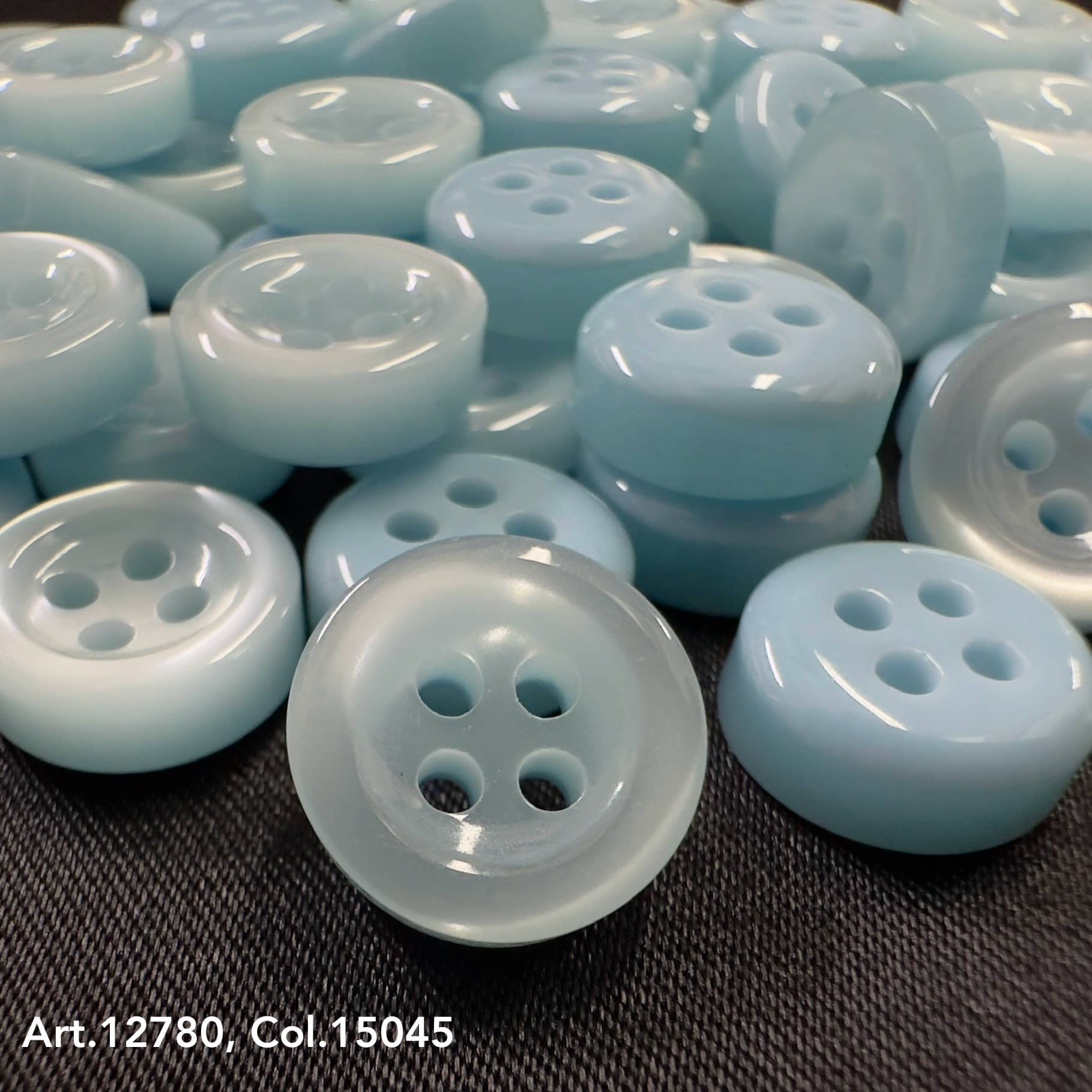 18L four hole plastic buttons in light blue color from Buttonology Inc spread on dark surface